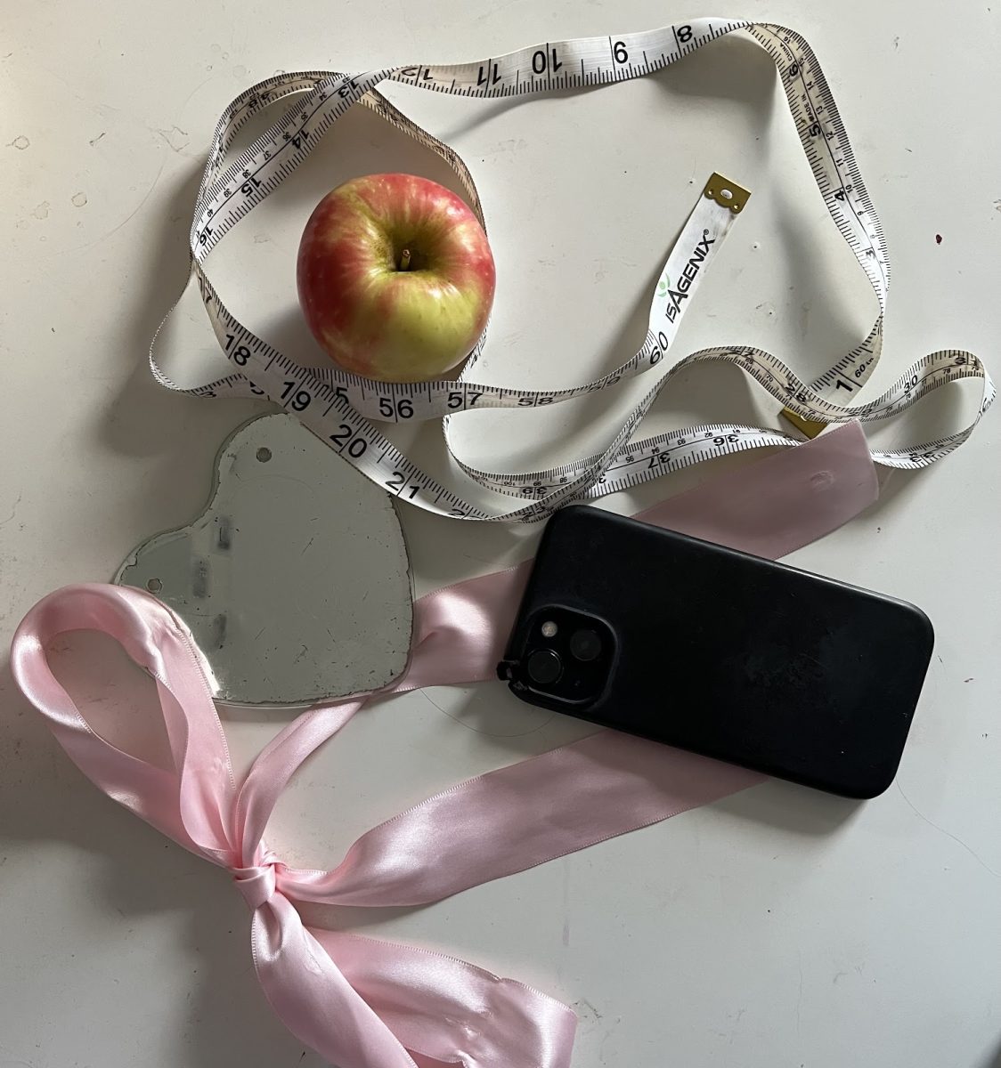 A phone, apple, mirror, ribbon, and measuring tape splayed across a table. Social media has increasingly become intertwined with most people’s lives, encouraging societal standards that push trending and unattainable body types. Photo by 