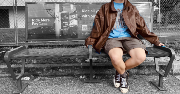 A person seated on a public bench at a bus stop in Portland, hands clasping bars dividing the bench into segments. The bars installed on these benches are a common form of hostile architecture, intended to prevent homeless people from lying down or resting at bus stops. Photo by Easton Atlansky.