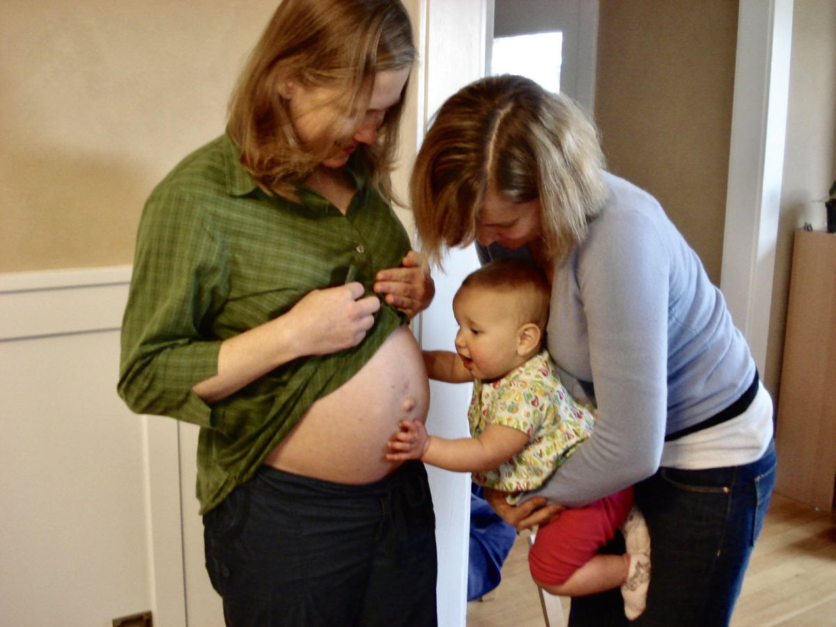 A pregnant woman lifts her shirt to allow a baby to touch her belly, while the baby is held by her own mother. Midwives have a unique role in providing support to pregnant people that goes beyond medical care. Photo by Joe MacKenzie. 